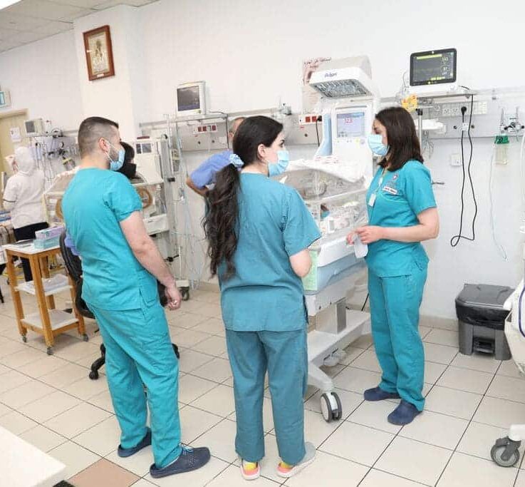 Three healthcare workers in scrubs and masks standing around a neonatal incubator in a hospital ward.