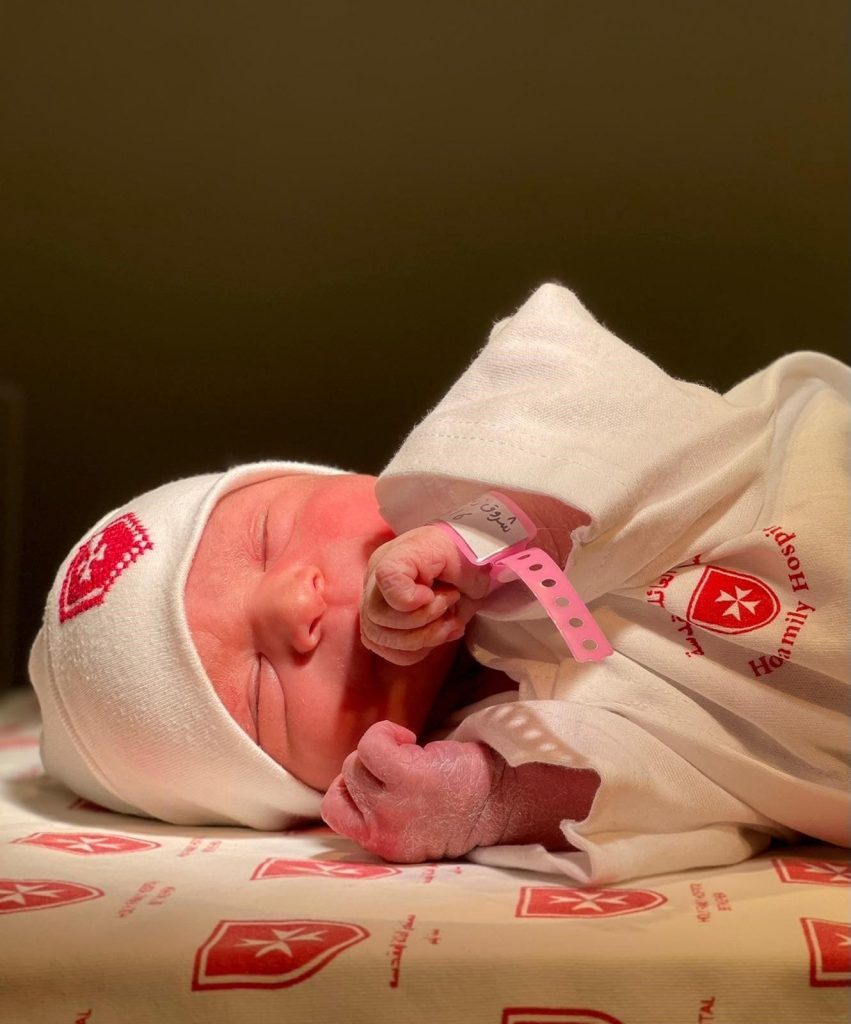 Newborn baby in hospital attire and hat, wearing an identification wristband, resting on a patterned blanket.