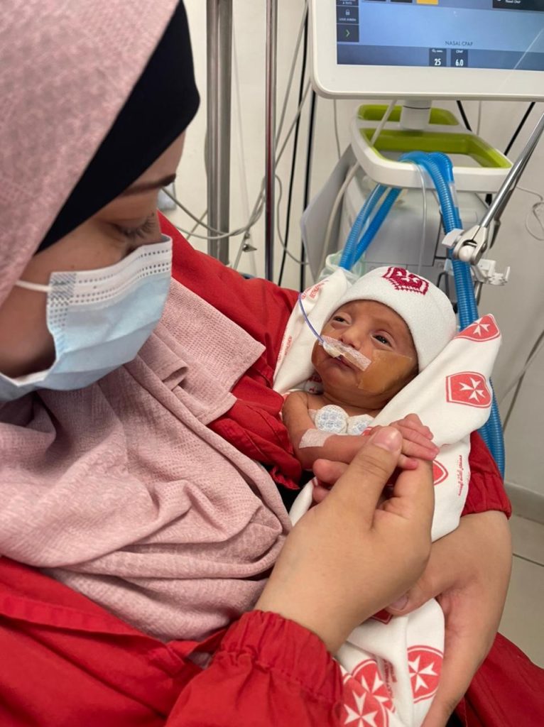 Woman wearing a mask holding a premature baby wrapped in a white blanket with a nasal tube.