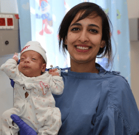 Smiling woman in medical attire holding a newborn baby in a hospital setting.