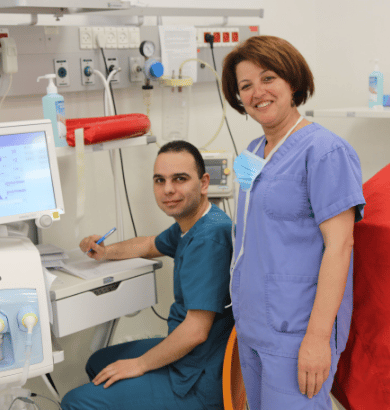 Two healthcare workers in scrubs smiling in a hospital setting, with one seated at a workstation.