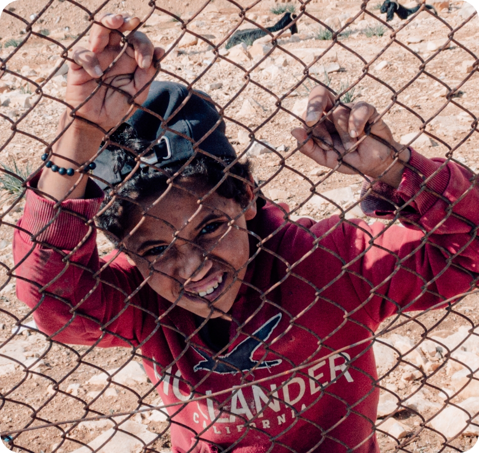 Smiling child in a red hoodie and backward cap holding onto a chain-link fence.