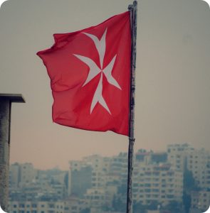Red flag with a white Maltese cross flying against a cityscape background.