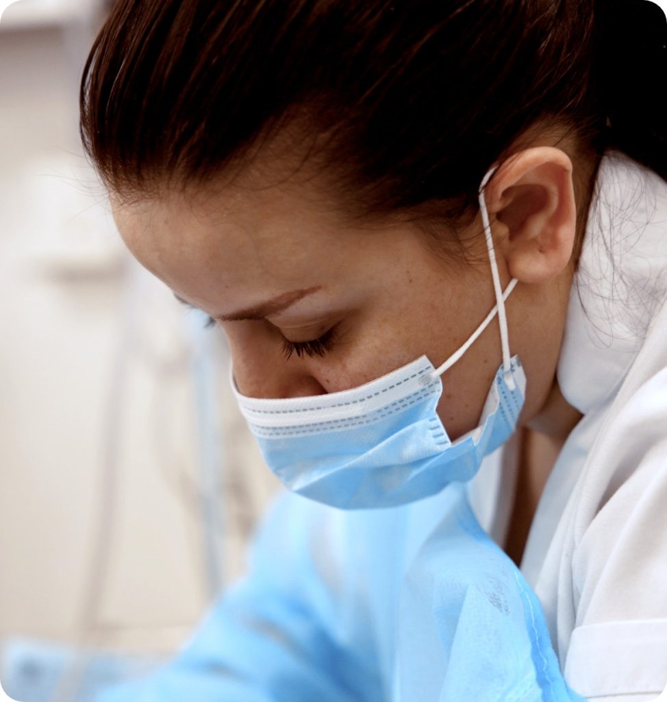 Healthcare worker wearing a face mask and blue protective gown, looking downward while working.