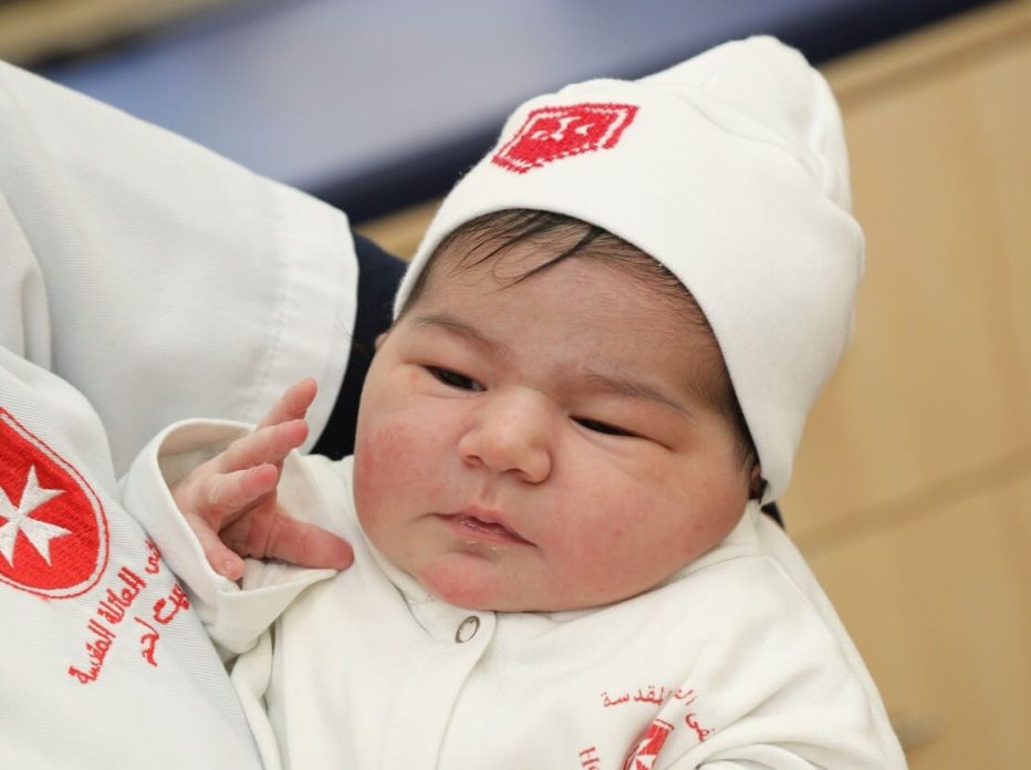 Newborn baby in white hospital outfit and hat with red emblem.