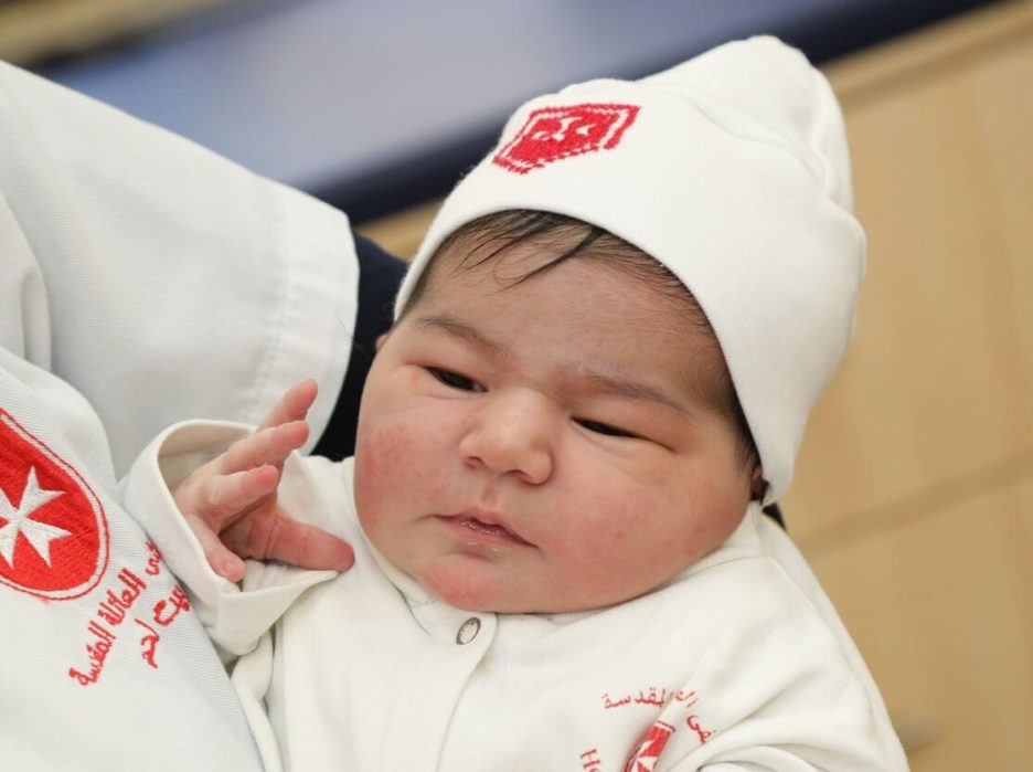 Newborn baby in white hospital outfit and hat with red emblem.