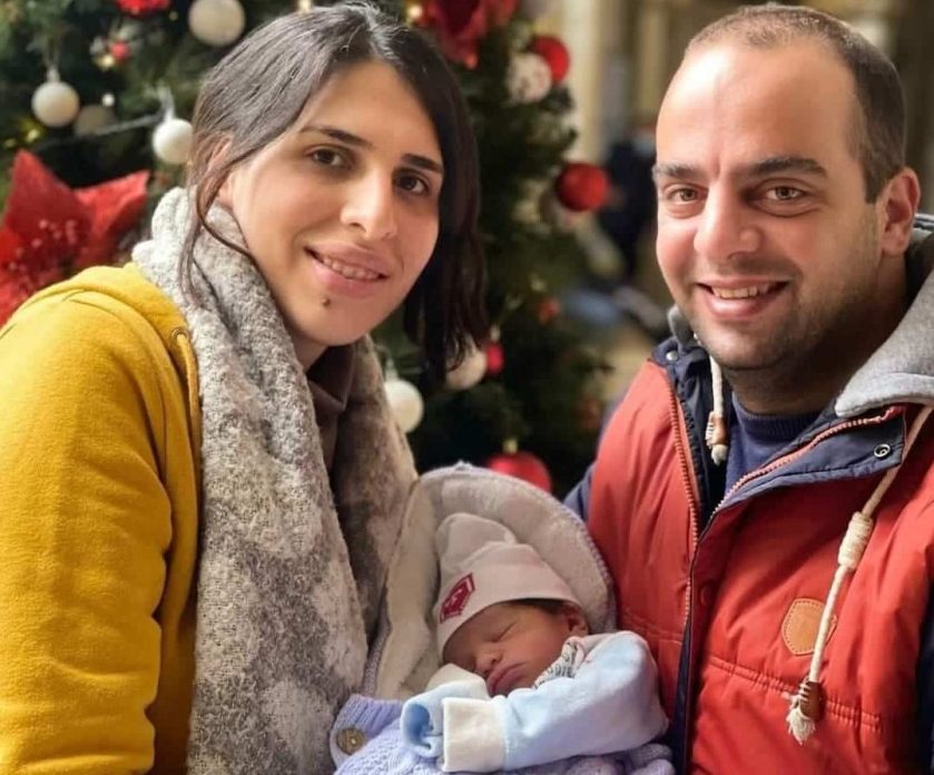 Smiling couple holding a newborn baby wrapped in a blanket, standing in front of a decorated Christmas tree.